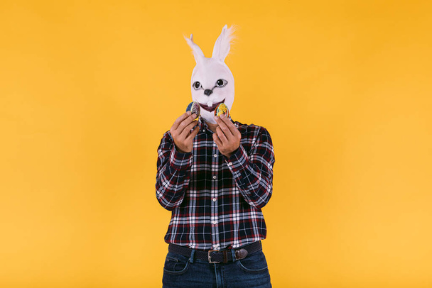 Zamaskowana osoba w masce króliczej w kratkę i dżinsach, trzymająca jajka wielkanocne na żółtym tle. Karnawał, przyjęcie, Wielkanoc i koncepcja świętowania. - Zdjęcie, obraz