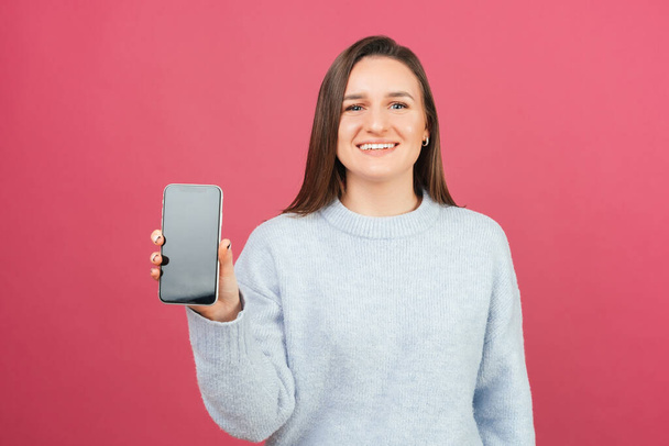 Jovem mulher sorrindo está segurando um telefone e mostrando tela para a câmera. - Foto, Imagem