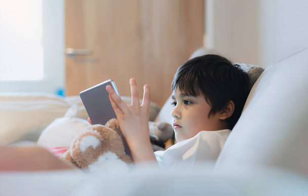 Cinematic portrait young boy playing game on tablet sitting on couch with sunlight shining from window,Kids sitting on sofa playing games online on internet, Childhood relaxing at home on weekend. - 写真・画像