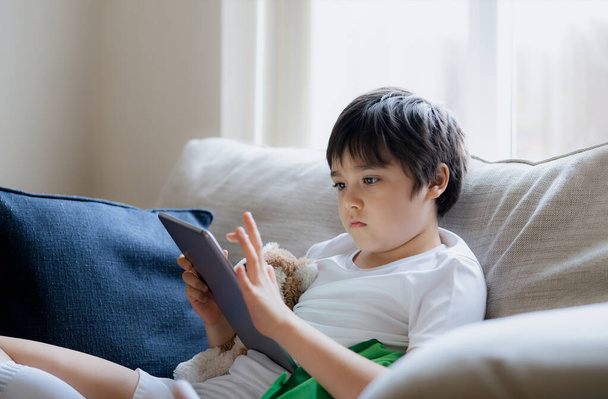 Młody chłopiec korzystający z tabletu gry w Internecie, Kid siedzi na kanapie oglądając lub rozmawiając z przyjacielem online, Dziecko relaksujące się w salonie rano, Dzieci z koncepcją nowej technologii - Zdjęcie, obraz