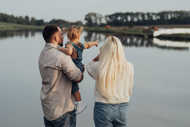 Back View of Young Family Standing Outdoor, Ο πατέρας κρατώντας το παιδί του στα χέρια, η μικρή κόρη δείχνει λίμνη στον μπαμπά και τη μαμά της - Φωτογραφία, εικόνα
