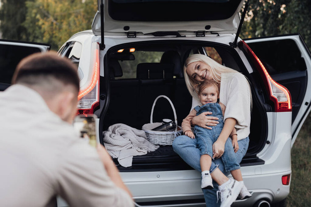 夫は、 SUV車のトランクを開けた中に座っている彼の妻と幼児の娘を撮影,ロードトリップ休暇の家族 - 写真・画像