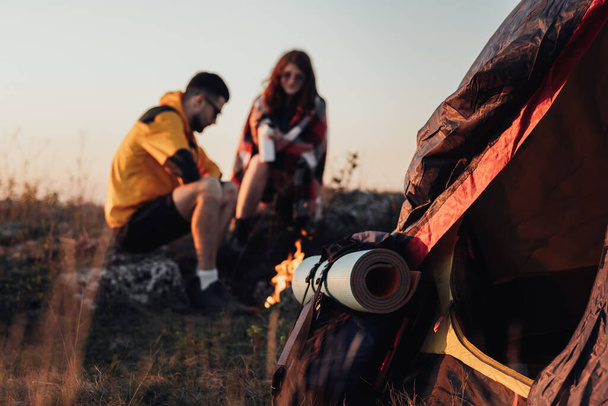 Zaino con stuoia e tenda a fuoco, sullo sfondo due giovani viaggiatori uomo e donna seduti vicino al caminetto durante il tramonto, concetto di viaggio - Foto, immagini