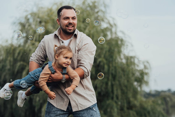 Happy Young Father Holding His Daughter in Arms and Imitating Flying and Playing with Bubbles Outdoor - Photo, Image