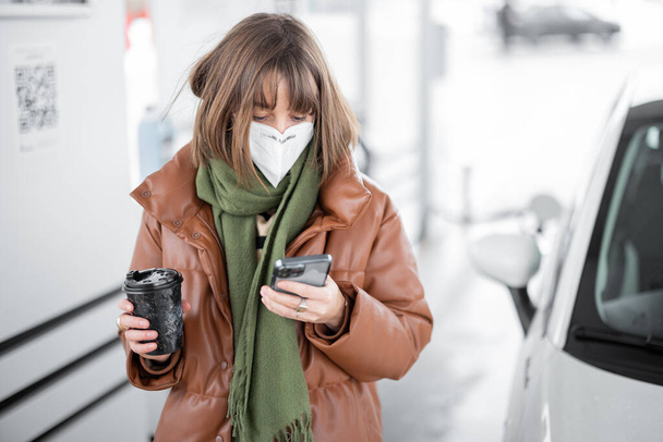 Woman in face mask holding smart phone and coffee cup while refueling car at gas station. - Photo, image