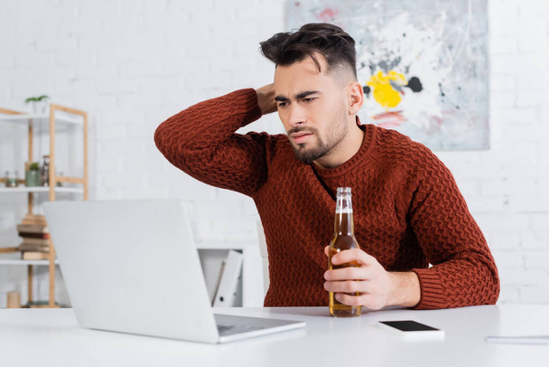 displeased bookmaker with bottle of beer looking at blurred laptop - Фото, изображение