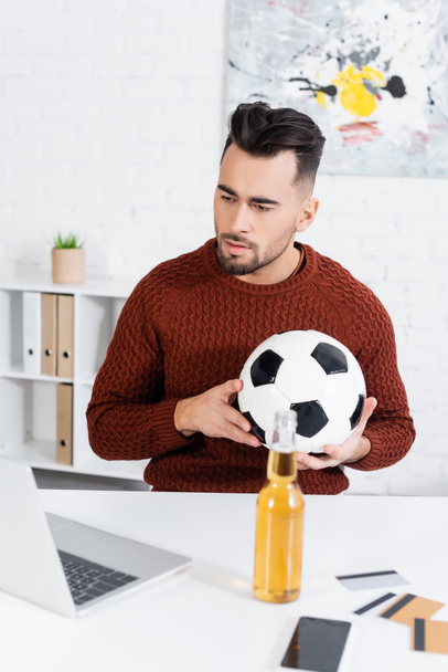concentrated bookmaker with soccer ball looking at laptop near bottle of beer and credit cards - 写真・画像