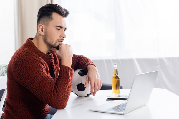 focused bookmaker with soccer ball looking at laptop near bottle of beer - Photo, Image