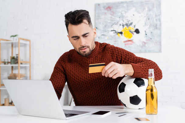 serious bookmaker holding credit card near soccer ball, laptop and beer - Fotó, kép