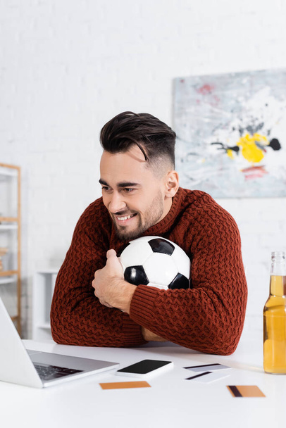 cheerful gambler hugging soccer ball near laptop, credit cards and beer - Fotografie, Obrázek