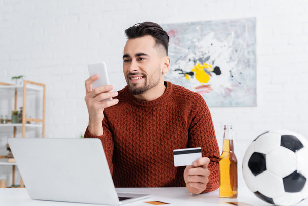 Lächelnder Spieler mit Kreditkarte blickt auf Smartphone in der Nähe von Laptop, Bier und Fußball - Foto, Bild