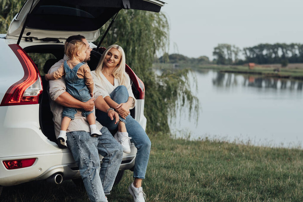 Junge Eltern mit kleiner Tochter sitzen im offenen Kofferraum des Autos, glückliche Familie genießt Roadtrip im Geländewagen - Foto, Bild