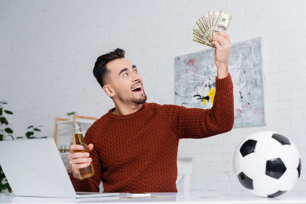 astonished gambler holding dollars and bottle of beer near laptop and soccer ball - Fotó, kép