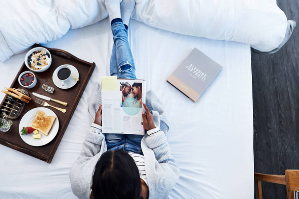 Breakfast in bed and some good books. High angle shot of an unrecognizable woman reading a magazine and having breakfast on her bed in her bedroom at home. - 写真・画像