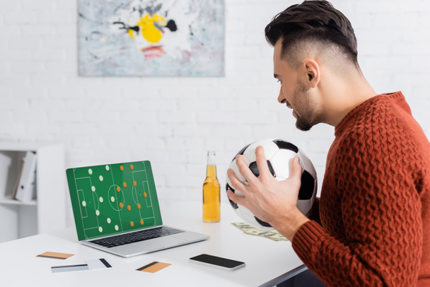 side view of bookmaker with soccer ball near credit cards and laptop with sports game strategy on screen  - Fotografie, Obrázek