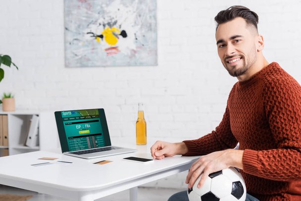 KYIV, UKRAINE - FEBRUARY 4, 2022: happy bookmaker with soccer ball looking at camera near laptop with sportsbet website - Zdjęcie, obraz