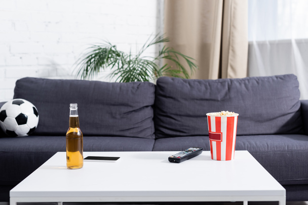 pöytä pullo olutta, TV kaukosäädin, ämpäri popcornia ja älypuhelin lähellä jalkapalloa sohvalla - Valokuva, kuva
