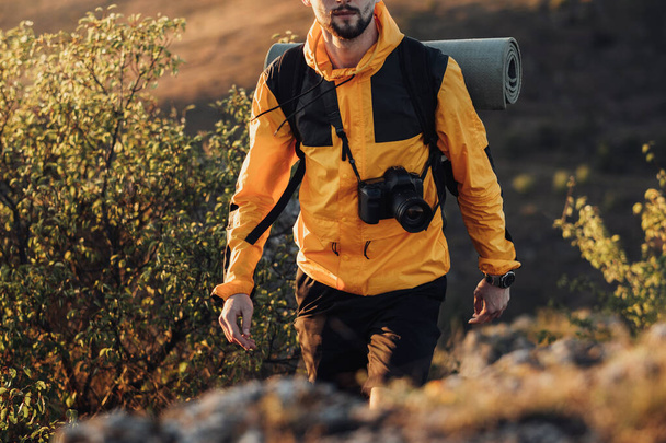 Nierozpoznawalny mężczyzna w stroju turysty Windbreaker, podróżnik z plecakiem i Camping Mat z aparatem cyfrowym Ciesz się swoją samotną wycieczką na wzgórzu podczas zachodu słońca - Zdjęcie, obraz