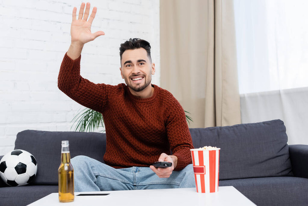 cheerful sports fan waving hand while watching game at home near soccer ball and beer - Zdjęcie, obraz