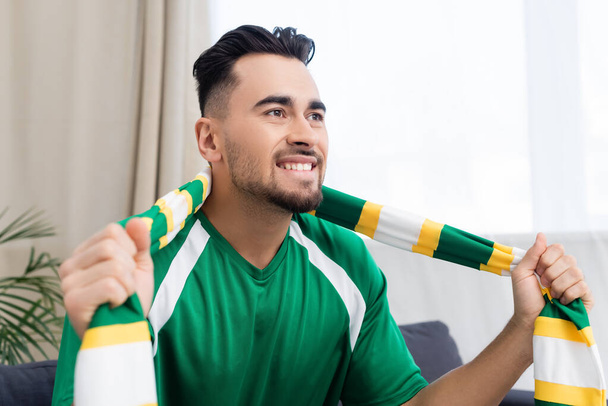 excited sports fan in striped scarf watching game at home - Photo, Image