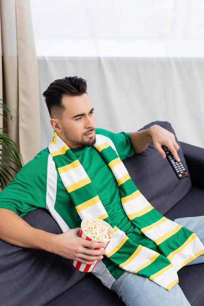 concentrated sports fan with tv remote controller and popcorn watching game at home - Fotografie, Obrázek