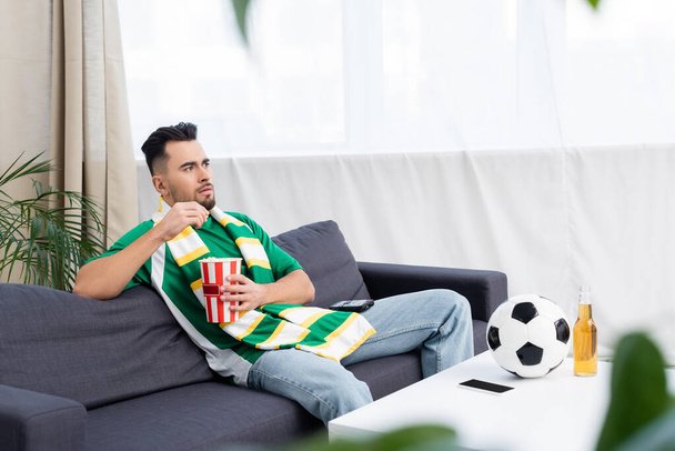 sports fan in striped scarf watching game on tv near soccer ball - Foto, imagen