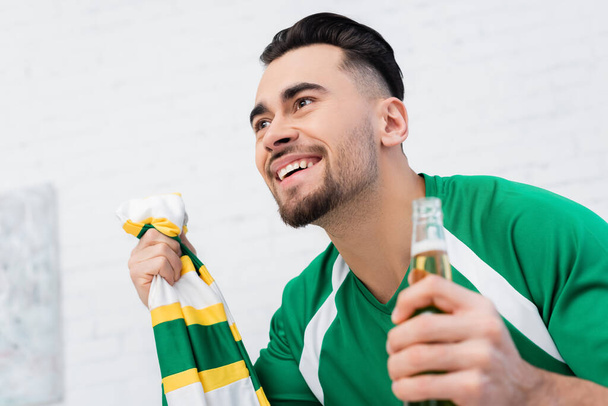 cheerful sports fan with striped scarf and beer watching match at home - Fotografie, Obrázek