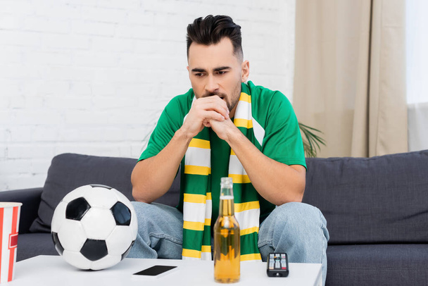 fanático de los deportes frustrados sentado cerca del mando a distancia de la televisión, pelota de fútbol y cerveza - Foto, Imagen
