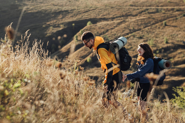 Travel Couple Man and Woman with Backpacks and Camping Mat Holding by Hands Moving on the Hill During Sunset - Photo, image