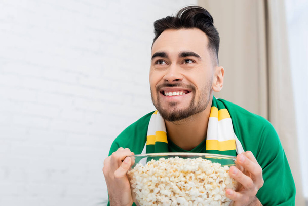 thrilled sports fan holding bowl of popcorn while watching championship at home - Photo, Image