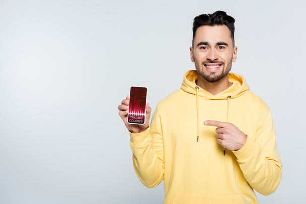 happy man pointing at smartphone with trading courses app isolated on grey - Fotoğraf, Görsel