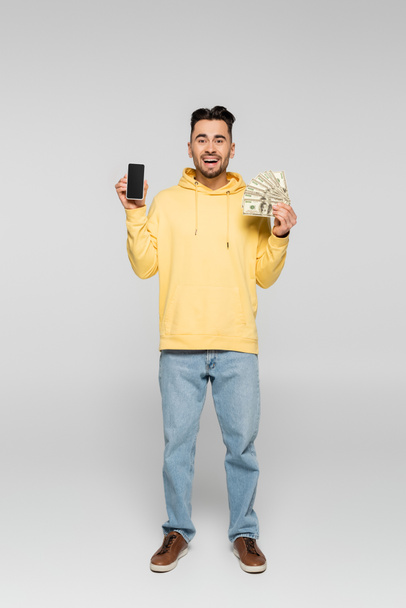 full length of smiling gambler with dollars and cellphone with blank screen on grey - Photo, Image