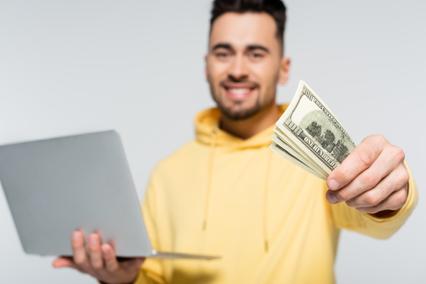 casa de apuestas borrosa que muestra billetes de dólar mientras sostiene el ordenador portátil sobre fondo borroso aislado en gris - Foto, imagen