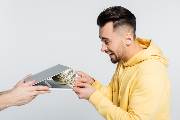side view of excited gambler holding money near laptop isolated on grey - Photo, image