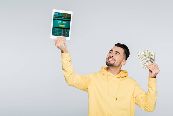 KYIV, UKRAINE - FEBRUARY 4, 2022: cheerful bookmaker holding dollars and digital tablet with sportsbet app isolated on grey - Φωτογραφία, εικόνα