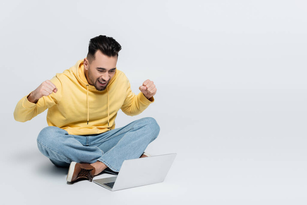 cheerful man showing triumph gesture while sitting with laptop on grey - Фото, зображення