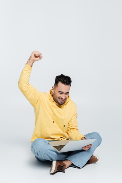 збуджений букмекер, що показує жест виграшу, сидячи з ноутбуком на сірому
 - Фото, зображення