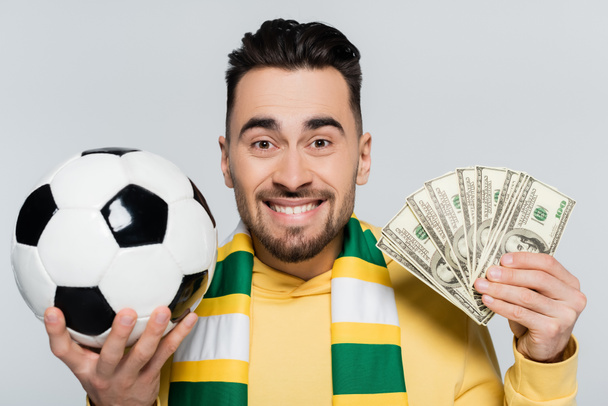 excited gambler with dollars and soccer ball looking at camera isolated on grey - Photo, Image