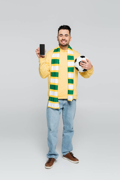 повна довжина щасливого гравця з футбольним м'ячем, що показує мобільний телефон з порожнім екраном на сірому
 - Фото, зображення