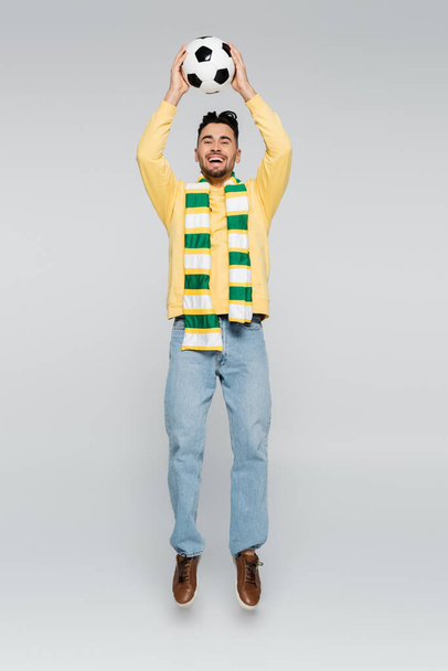 full length of overjoyed sports fan jumping with soccer ball in raised hands on grey - Foto, immagini