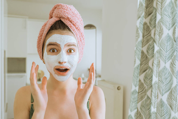 dospívající dívka s ručníkem omotaným kolem mokrých vlasů, s bílou hliněnou kosmetickou maskou na obličeji, se dívá do kamery s překvapením nebo potěšením. Kosmetický zákrok doma. - Fotografie, Obrázek