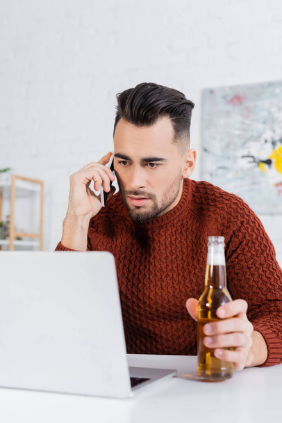 serious bookmaker with bottle of beer talking on smartphone near laptop - 写真・画像