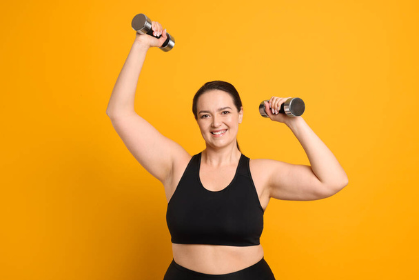 Happy overweight woman doing exercise with dumbbells on orange background - Photo, Image