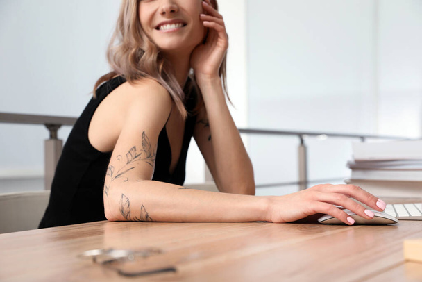 Beautiful woman with tattoos on arms using computer at table indoors, closeup - Fotó, kép