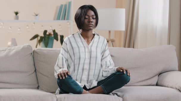Fiatal, fókuszált afro-amerikai nő ül lótuszpozícióban, csukott szemmel egészséges, nyugodt lány lazít a kanapén, lenyugszik, mély levegőt vesz, meditál, otthon pihent, megszabadulva a stressztől. - Felvétel, videó