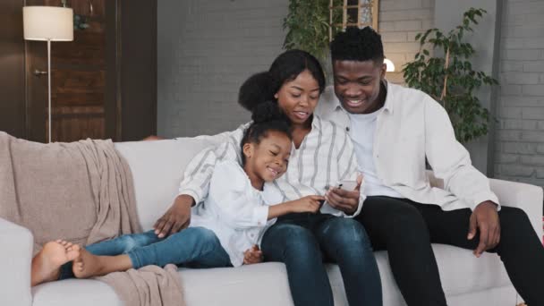 Afrikaanse familie praten lachen kijken naar smartphone met behulp van grappige apps zitten op de bank kiezen van online winkelen met telefoon glimlachende moeder vader en dochter ontspannen thuis mobiele telefoon online kijken video - Video