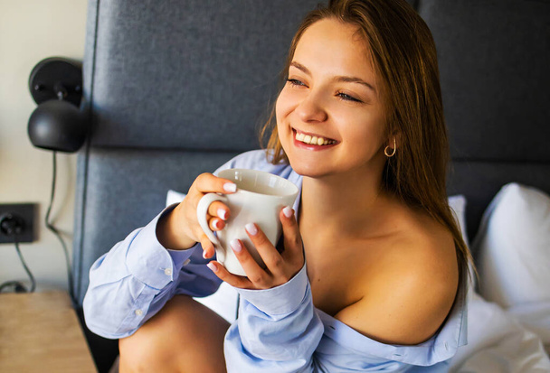 Image of beautiful young woman in blue shirt smiling and drinking tea on bed in white apartment - Valokuva, kuva