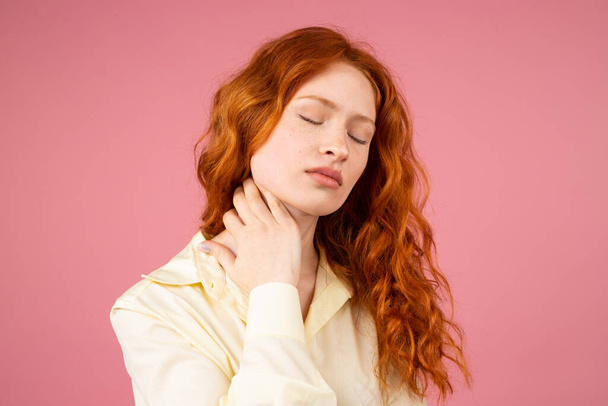 Close up photo of a redhead woman posing with closed eyes and keeping her hand on her neack on pink background. - Photo, Image