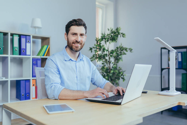 Happy and successful man working on laptop in home office, sitting at table in bright room, smiling looking at camera - Photo, Image