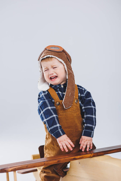 Мальчик плачет в кожаной куртке и пилотской шляпе, деревянном самолете, коричневых чемоданах. Детские деревянные игрушки. Детские эмоции - Фото, изображение